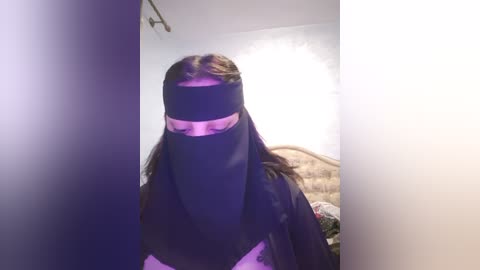 Media: A video of a person wearing a black niqab, with long brown hair visible, standing in a dimly lit room with white walls and a bed in the background.