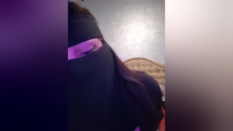 Media: A video captures a close-up of a person wearing a black mask, with purple lighting highlighting the mask's texture and the individual's eye. The background shows a dimly lit, beige wall with a wooden headboard. The image conveys a sense of mystery and intrigue.