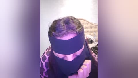 Media: Video of a person in a black mask and a polka-dot shirt, with long brown hair, standing in a dimly lit bedroom with a bed and wall art in the background.