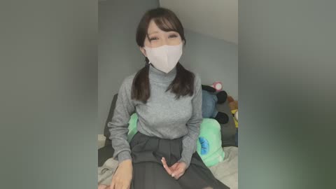 Media: Video of an Asian woman with straight, shoulder-length brown hair, wearing a white mask, gray turtleneck, and a black skirt, seated on a bed with plush toys, in a dimly lit room.