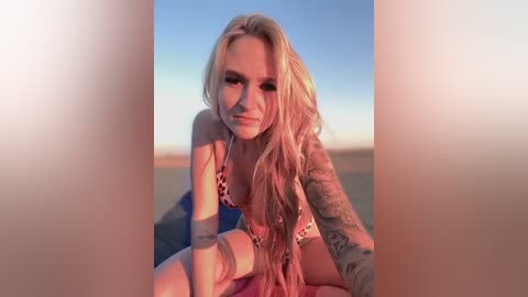 Media: Video of a young blonde woman with long hair and a tattooed arm, wearing a leopard-print bikini, sitting on a beach at sunset, background blurred.