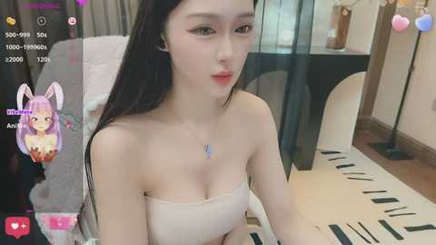 Media: Video of a young Asian woman with long black hair, fair skin, and light pink lipstick, wearing a strapless beige top, in a modern, brightly lit room.