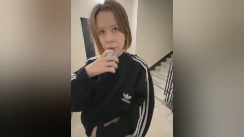 Media: A video of a young girl with light skin and shoulder-length brown hair, wearing a black Adidas jacket and blue nail polish, standing in a dimly lit hallway.