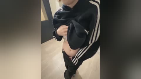 Media: A video of a person lifting a black Adidas jacket to reveal their bare midriff and partially lowered pants, standing in a beige-tiled hallway with a dark blue wall.