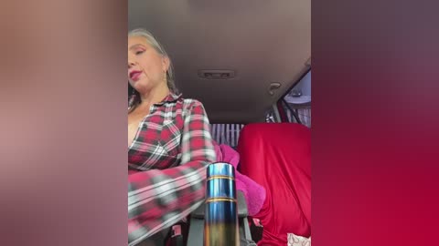 Media: Video of a blonde woman with medium-length hair, wearing a red plaid shirt, leaning back in a red seat, with a bottle in her hand.