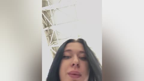 Media: A video of a young woman with long, straight black hair, fair skin, and a neutral expression, taken from a low angle. The background features a white, industrial ceiling with exposed beams and a large, square window.