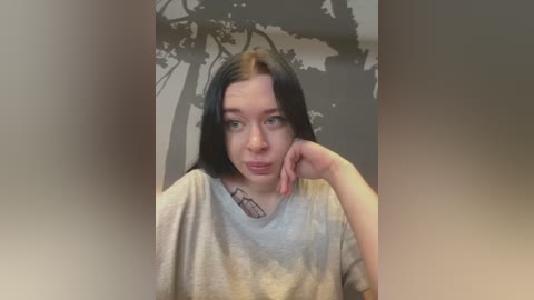 Media: A video of a young Caucasian woman with long black hair, light skin, and a grey T-shirt, leaning on her elbow, with a wall mural featuring trees in the background.