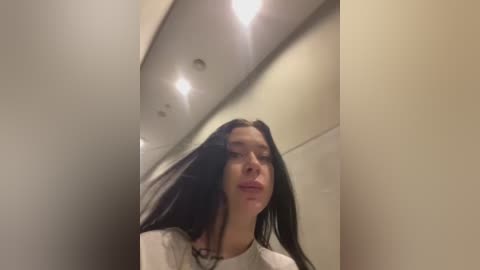 Media: Video of a young woman with long black hair, fair skin, and a neutral expression, taken indoors with white walls and recessed lights.