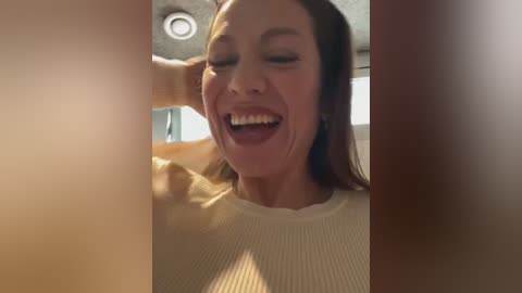 Media: Video of a woman with fair skin and long brown hair, wearing a beige sweater, laughing joyfully in a dimly lit room with blurred background.