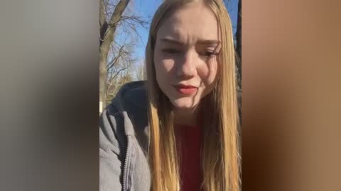 Media: Video of a young, fair-skinned, blonde-haired woman with a neutral expression, wearing a red top and gray hoodie, standing in a park with bare trees under a clear blue sky.