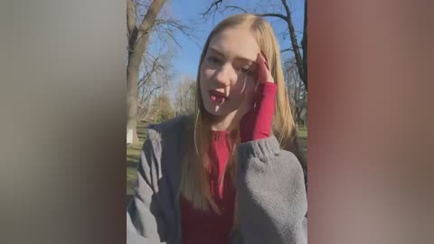 Media: A video of a blonde woman with fair skin, wearing a red top and gray cardigan, standing outdoors in a park, with bare trees and blue sky in the background.