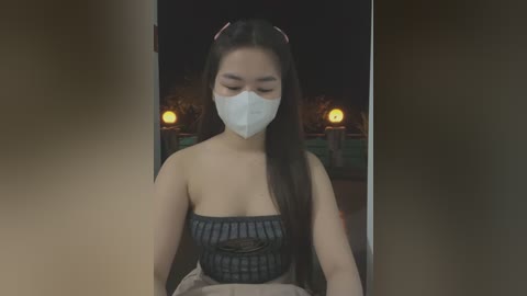 Media: Video of an Asian woman with long black hair and light skin, wearing a black lace-up top, white mask, and red headband. Background shows dimly lit outdoor scene with two yellow lights.