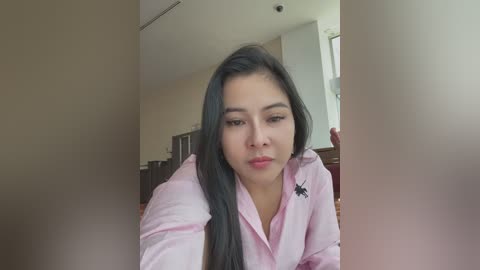 Media: Video of a young woman with long, straight black hair, wearing a light pink shirt with a spider pin, indoors, looking pensive, with blurred background.