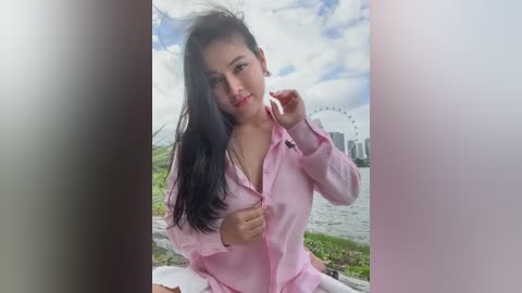 Media: Video of a young Asian woman with long black hair, wearing a pink button-up shirt, standing outdoors by a lake with a Ferris wheel in the background.