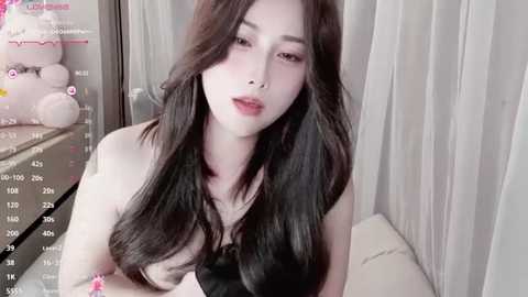 Media: A video of an East Asian woman with long black hair, pale skin, and full lips, wearing a black top. She is indoors, near a window with white curtains.