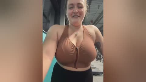 Media: Video of a smiling, plus-sized woman with fair skin and auburn hair, wearing a brown sports bra and black leggings, standing indoors with industrial background.