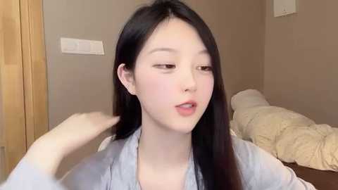 Media: A video of an East Asian woman with long black hair, fair skin, and a slender physique, wearing a light blue shirt, adjusting her hair in a cozy, beige-toned bedroom.