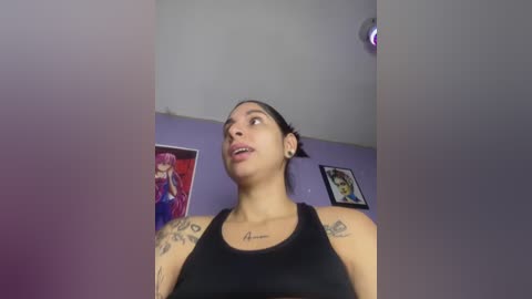 Media: Video of a Latina woman with medium skin tone, dark hair tied back, wearing a black tank top, tattoos on her arms. She stands in a room with purple walls, posters of anime characters, and a ceiling light.