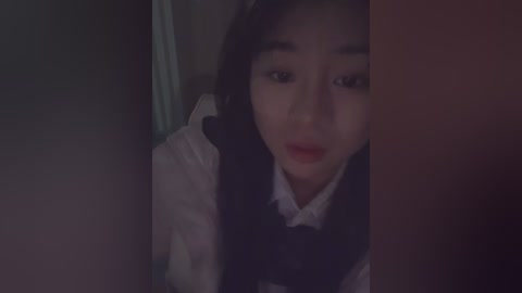 Media: A dimly lit video of a young Asian woman with dark hair and pale skin, wearing a white shirt and dark vest, looking concerned, taken from a low angle.