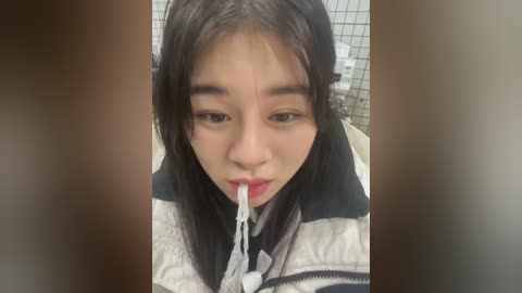 Media: Video of a young Asian woman with straight black hair, wearing a striped jacket, licking a white lollipop in a tiled bathroom.