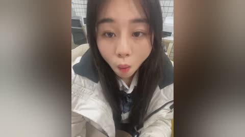 Media: Video of an East Asian woman with long black hair, fair skin, and a neutral expression, wearing a white jacket and a navy blue tie, seated in a bathroom with white-tiled walls and a green trash can in the background.