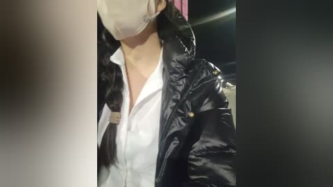 Media: A video shows a woman with long black hair, wearing a white surgical mask, a white button-up shirt, and a black glossy jacket. The background is blurred, indicating an indoor setting.