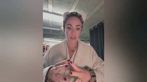 Media: Video of a young woman with fair skin, brown hair, and dark eyebrows, wearing a beige robe with red nails, standing in a dimly lit room with a textured ceiling and dark curtains.