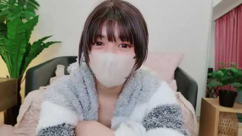 Media: Video of a young woman with short dark hair, wearing a white mask and a gray and white fuzzy blanket, sitting on a bed with a green plant and pink curtains in the background.