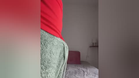 Media: A video showing a person's lower body in a red shirt, wearing a green towel, standing next to a bed with white sheets and a pink pillow in a simple, minimalist room.