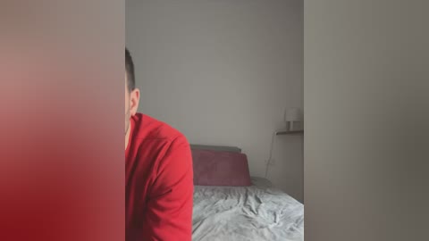 Media: A video of a man in a red shirt standing in a minimalist bedroom with a gray bed, white walls, and a nightstand.