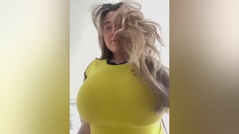 Media: Video of a woman with long blonde hair, large breasts, wearing a tight yellow shirt, standing against a white background.