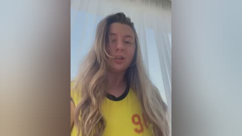 Media: A video of a young woman with long, wavy blonde hair wearing a yellow T-shirt, standing in a softly lit room with white curtains in the background.