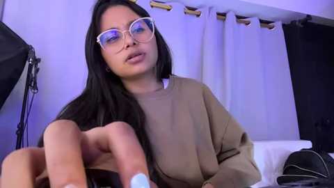 Media: Video of a young woman with long black hair, wearing glasses, a beige sweater, and a white shirt, sitting in a dimly lit room with white curtains and a camera setup in the background.