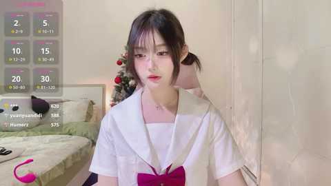 Media: Video of a young Asian woman with pale skin, dark hair, and red lipstick, wearing a white sailor uniform, standing in a bedroom with a bed and Christmas decorations.