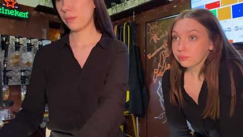 Media: Video of two women in a bar, one with long brown hair, black top, and pouty expression, the other with straight brown hair, black top, and playful pose.