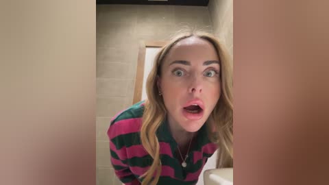 Media: A video of a blonde woman with wide eyes and open mouth, wearing a pink and green striped sweater, in a beige-tiled bathroom.