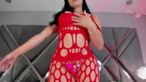 Media: Video of a curvaceous woman with long black hair, wearing a red, fishnet bodysuit, exposing her breasts and genitals. She is standing in a modern, grey-tiled room with geometric wall art.