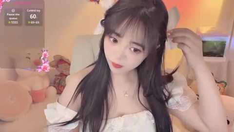 A video of a young Asian woman with long black hair, fair skin, and large eyes, wearing a white off-shoulder dress, adjusting her hair in a bedroom with plush toys and a colorful, cheerful background.