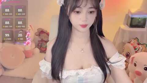 A video of an East Asian woman with long black hair, wearing a white off-shoulder dress, in a cozy room with plush toys and a temperature display.