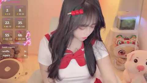 Video of a young East Asian woman with long black hair, wearing a red and white sailor outfit, playing a video game. Background features a pink plush toy and a monitor.