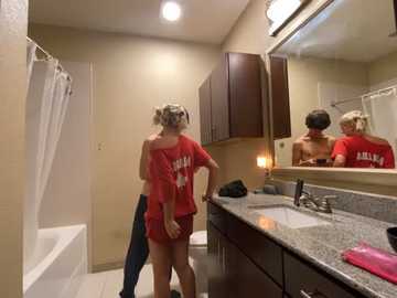 Media: Video of a blonde woman in a red off-shoulder shirt and shorts standing in a bathroom with beige walls, a granite countertop, and a mirror.