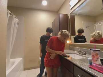 Media: Video of a bathroom with two young people; a blonde girl in a red dress, a boy in a black t-shirt and baseball cap, using a smartphone.