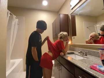 Media: Video of a young couple in a bathroom: man in black shirt, lifting woman's red shorts, revealing her buttocks; granite countertop, mirror, and white bathtub in background.