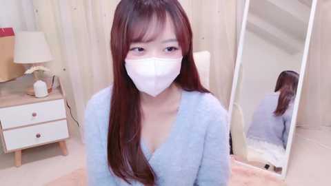 Media: Video of an East Asian woman with long brown hair, wearing a light blue sweater, white mask, and a white t-shirt, sitting in a cozy, softly lit bedroom with a wooden nightstand, mirror, and white curtains.