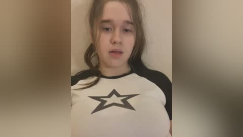 Media: Video of a young woman with fair skin and long brown hair, wearing a black-and-white star-patterned T-shirt, standing against a blurred beige background.