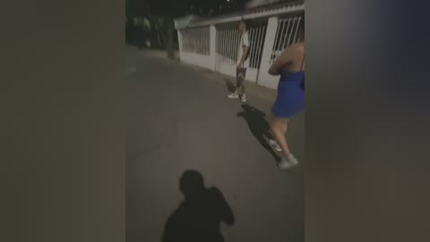 Media: Video of a dimly lit, narrow street at night. A woman in a blue dress and white sneakers rides a skateboard, followed by a man in dark clothing. The scene is captured from a low angle, with a blurred shadow in the foreground.