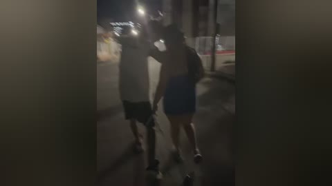 Media: A dimly lit video captures a man and woman walking on a dark, narrow street, holding hands. The woman wears a blue dress and a hat, while the man is in a black shirt and shorts. Streetlights illuminate the scene, casting shadows.