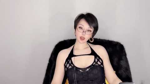 Media: Video of a young, pale-skinned woman with short black hair, wearing glasses, a black halter top, and sitting on a black fur chair against a plain white background.