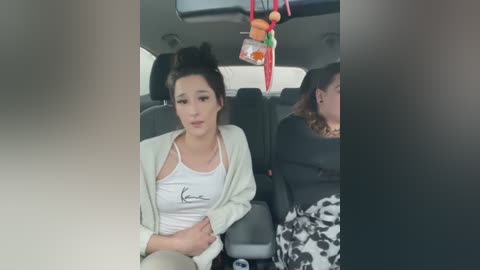 Media: Video of two women in a car, one with dark hair in a messy bun, wearing a white top and beige jacket, the other with long hair, wearing a black top.