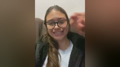 Media: Video of a young woman with long brown hair, wearing glasses and a black cardigan over a white shirt, smiling, with a blurred background.